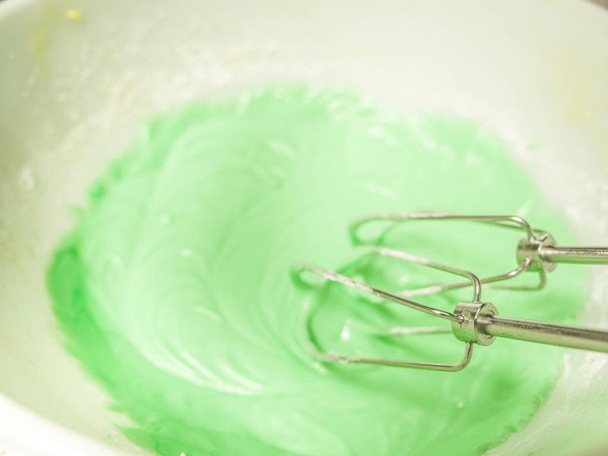 Close up view of pastel or mint green colored frosting and metal beaters in a white ceramic mixing bowl sitting on top of a kitchen counter. - Foto, Bild