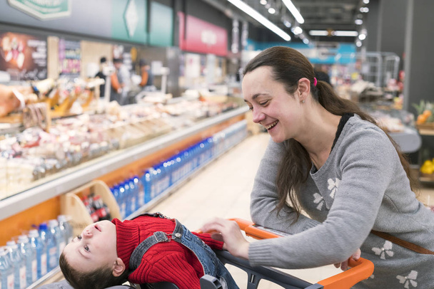 Μια νεαρή γυναίκα με ένα μικρό γιο σε ένα σούπερ μάρκετ. Το αγόρι κάθεται σε ένα καλάθι μωρού και γέλια - Φωτογραφία, εικόνα
