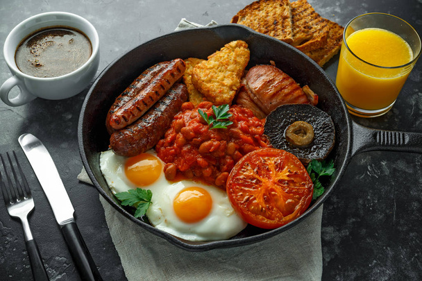 Kompletní anglická snídaně s slaninou, klobása, sázené vejce, pečené fazole, opékané brambory a houby v rustikálním pánev, pan. Cup káva, pomerančový džus - Fotografie, Obrázek