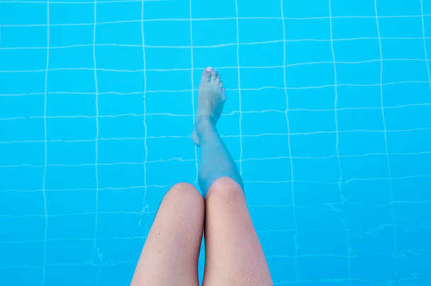 女の子は、澄んだ青い水のプールで彼女の足を跳ねます。女性は夏の暑い日にプールサイドで休んでいます。女性の脚線美。女性の足と水の足. - 写真・画像