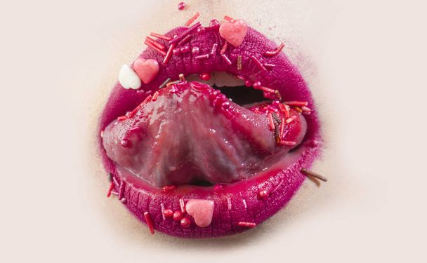 Tong in open vrouwelijke mond. Zoete sexy lippen met snoep. Vrouw likt roze harten. Verleidelijke sensuele mond van jong meisje met roze lippenstift, zoete cosmetica concept. Heerlijk dessert en erotische voedsel - Foto, afbeelding