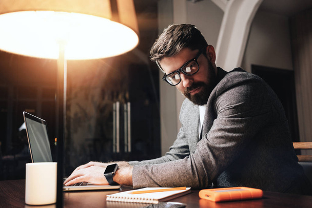 Bearded businessman in eye glasses working by the contemporary laptop at night coworker studio. Young project manager typing text on notebook. Film effect - Φωτογραφία, εικόνα