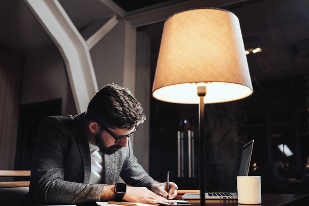 Creative businessman working with paper documents and contemporary laptop at late evening in loft office. Bearded young man sits by the wooden table with lamp and notebook. Creative manager write new plan - Φωτογραφία, εικόνα