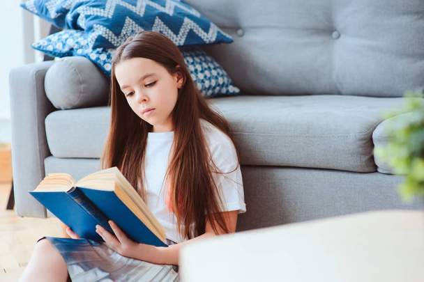 smart kid girl reading interesting book  at home, sitting with couch on background. Learning and education concept - 写真・画像