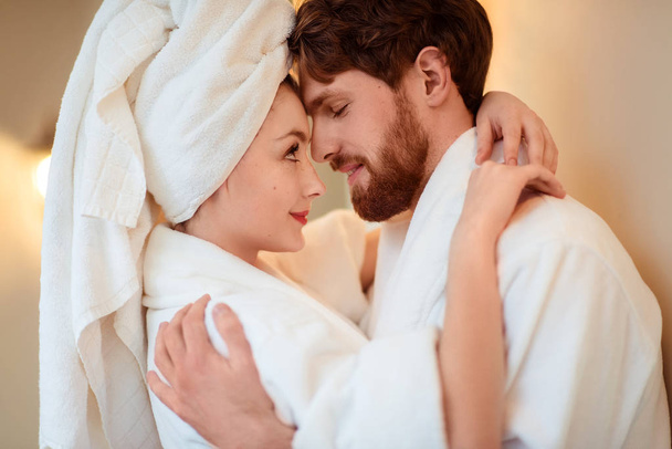 Close up shot of beautiful woman and her husband cuddle each other, express love, wear comfortable bathrobes, relax after taking shower or having spa procedure in luxury hotel. Beauty and relations - Фото, изображение