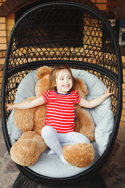 little girl sitting on chair on the terrace with toy bear. - Φωτογραφία, εικόνα