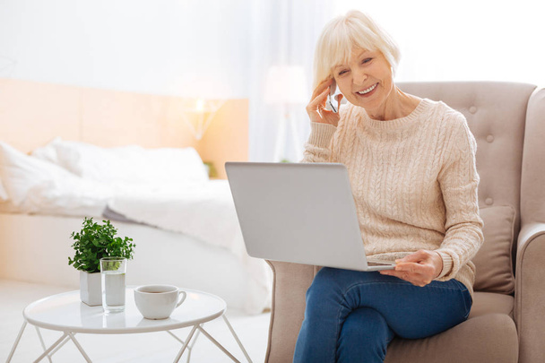 Emotional smart pensioner talking on the phone while sitting with a laptop - Valokuva, kuva