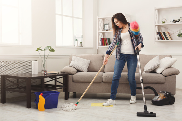 Geschokt vrouw reinigen van huis met tal van instrumenten - Foto, afbeelding