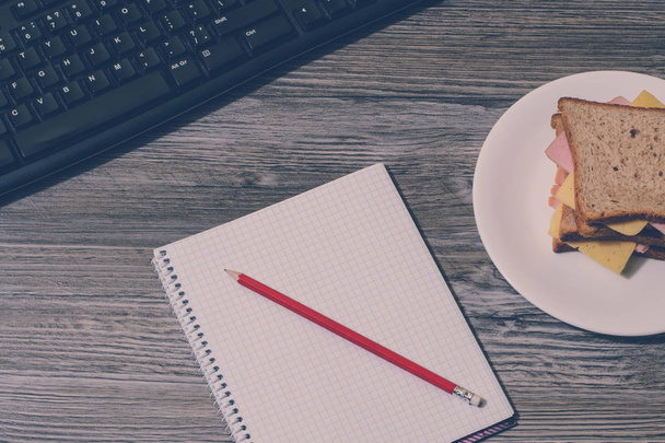 Breken op het werk. Lekkere kaas sandwich, notitieboekje en potlood met toetsenbord op grijze houten achtergrond. Accent op een laptop. Verticale weergave, vintage effect - Foto, afbeelding
