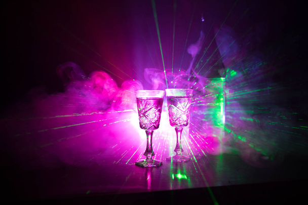 Twee glazen wodka met fles op donker mistige club stijl achtergrond met gloeiende lichten (Laser, Stobe) Multi gekleurd. Club drinkt themadecoratie. Lege ruimte - Foto, afbeelding