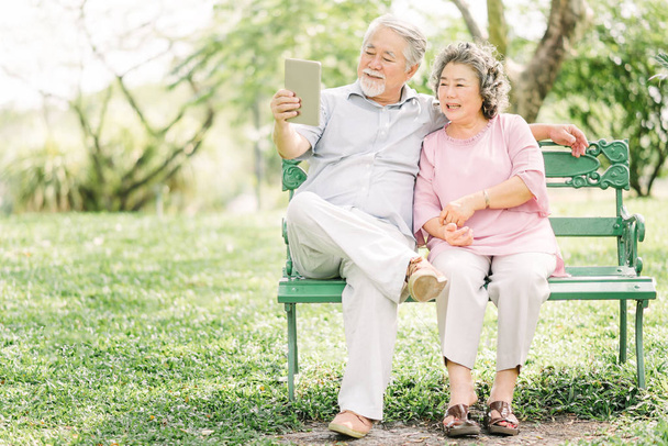 Šťastný starší Asijské pár sedí na lavičce společně pomocí digitálních tabletu venkovní  - Fotografie, Obrázek