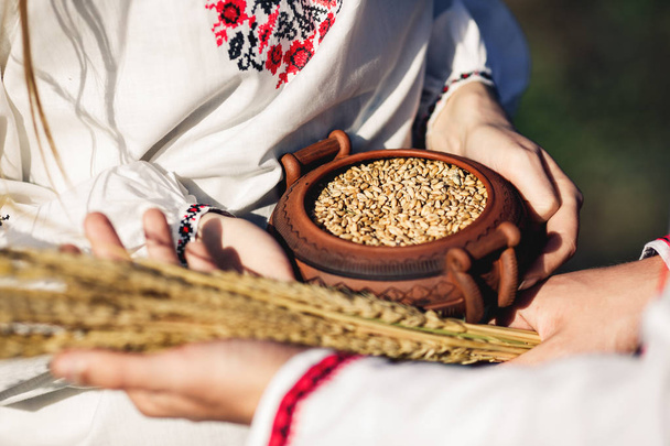 Espiguillas de trigo y una olla de semillas de trigo se llevan a cabo en las manos de un chico y una niña en camisas bordadas
 - Foto, Imagen