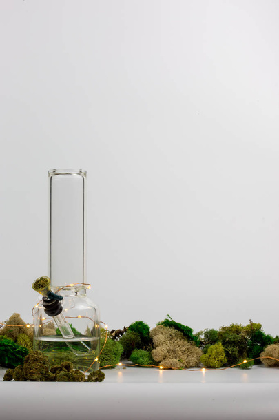 Скляний бонг, наповнений бур'янами, оточений бутонами бур'янів, різноманітним мохом і струнними вогнями проти білого
 - Фото, зображення