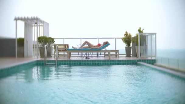 Eine Frau im rosafarbenen Bikini genießt den Blick auf das Meer am Pool auf dem Dach, das Mädchen sonnt sich auf einer Liege. 4k, Zeitlupe - Filmmaterial, Video