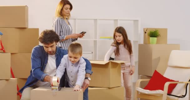 Good looking father with a little son packing a box with a sticky tape and a mother with a teen daughter on the background. Moving out the house. Indoors - Footage, Video