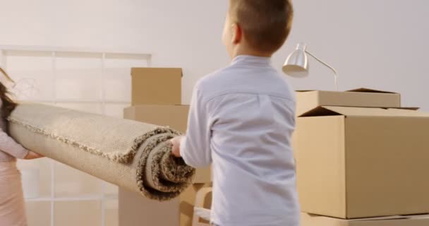 Pequeños niños encantadores, hermano y hermana, trayendo una alfombra grande a la sala de estar llena de cajas llenas durante la mudanza en el nuevo hogar. De interior
 - Metraje, vídeo