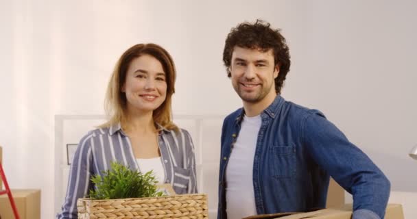 Good looking married couple looking at each other and then to the camera while moving in the new apartment and holding plant and box in hands. Indoors. Portrait. close up. - Imágenes, Vídeo