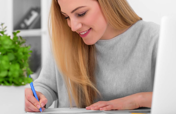 Aantrekkelijke jonge vrouw in een goed humeur werkt met documenten in het kantoor of thuis. Schrijft, papieren aan de balie vult met een laptop. - Foto, afbeelding