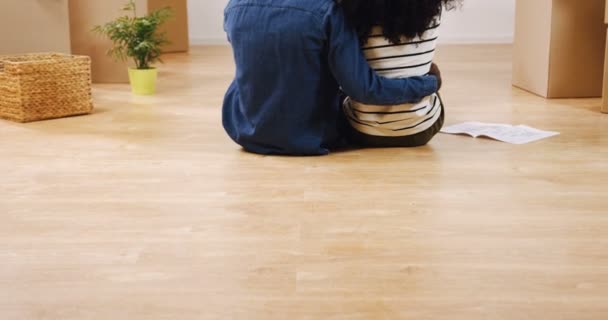 Задний вид на обнимающего афроамериканского парня и девушку, сидящих на полу в середине комнаты, полной коробок, и они смотрят на план квартиры, думая о дизайне для
 - Кадры, видео