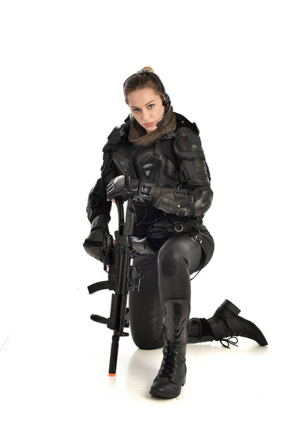 ホワイト スタジオの背景に分離された銃を保持しての座りポーズ、黒戦術的なアーマーを着ていた女性の兵士の完全な長さの肖像画. - 写真・画像