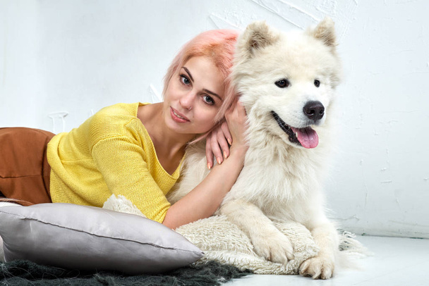 attractive young girl with her pet Samoyed posing lying on the white floor. Studio portrait with gentle warm tones. - Photo, image