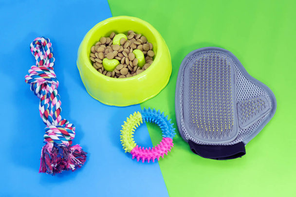 Pet supplies about bowl with dry food, rubber toys, glove brush and rope on blue and green background. - Photo, Image