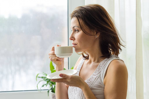 Donna matura beve caffè del mattino e guarda fuori dalla finestra a casa. Foto di profilo femminile grave
 - Foto, immagini