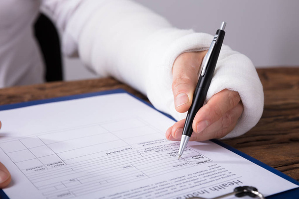 Close-up Of A Businessperson With Broken Arm Filling Health Insurance Claim Form On Wooden Desk - 写真・画像