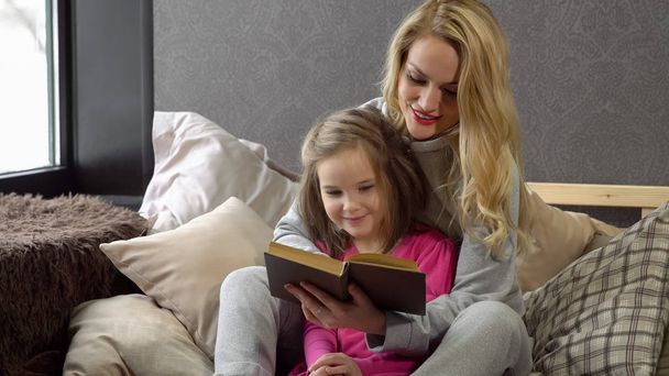 Mother and daughter sitting on the bed in an embrace and reading a book - Photo, image