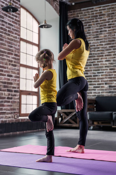 Madre e hija con ropa deportiva practicando yoga juntas meditando de pie sobre una pierna con las manos en posición de oración en el apartamento loft
 - Foto, imagen