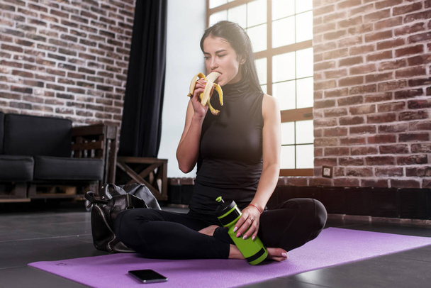 Portrait de jeune femme caucasienne se reposant après l'entraînement tenant une bouteille de sport assise sur un tapis avec les jambes croisées dans la salle de gym avec mezzanine intérieur
 - Photo, image