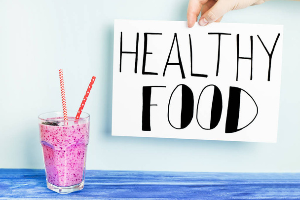 Banana currant smoothies of purple color with red straws stand on a wooden table on a blue background with a sign "Healthy food", which is held by the hand - Fotoğraf, Görsel