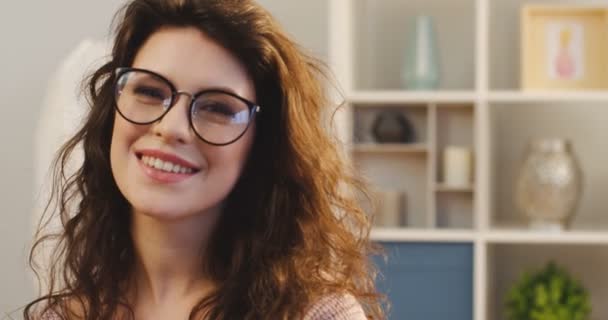 Close up of the attractive curly young woman in glasses smiling sincerely to the camera in the modern room of her apartment. Inside. Portrait - Πλάνα, βίντεο