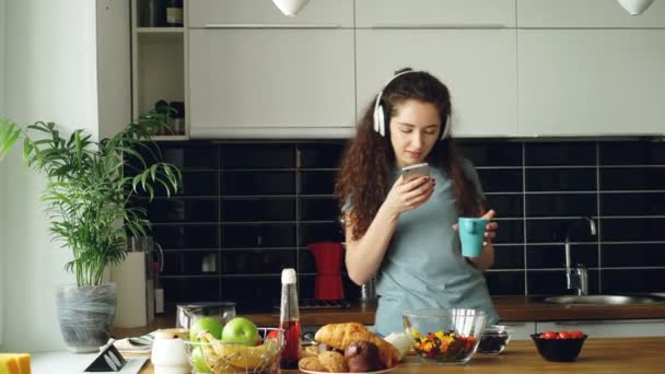 Atraktivní šťastná dívka tančí a zpívá v kuchyni při používání smartphone a poslech hudby doma ráno - Záběry, video