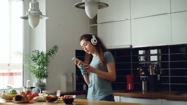 Attractive happy girl dancing and singing in kitchen while using smartphone and listening to music at home in the morning - Felvétel, videó
