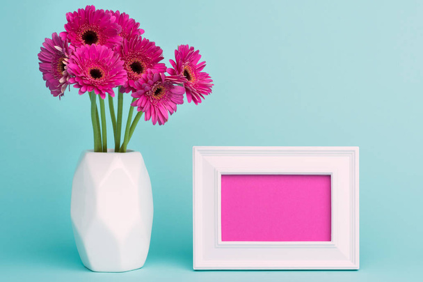 Happy Mother's Day, Women's Day, Valentine's Day or Birthday Pastel Candy Blue Coloured Background. Dark pink gerberas in a vase on a table with empty picture frame greeting card. - Photo, image