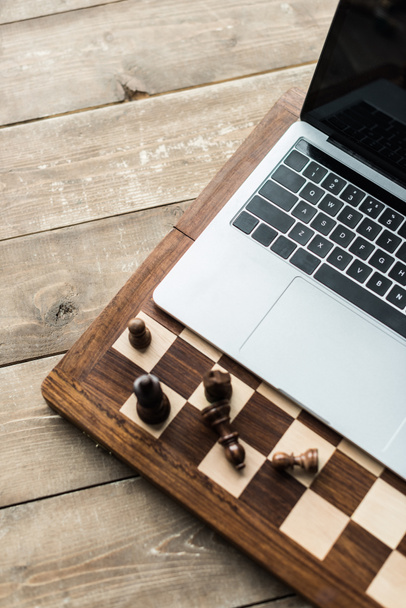 abgeschnittenes Bild eines Schachbretts mit Schachfiguren und Laptop auf rustikaler Holzoberfläche - Foto, Bild