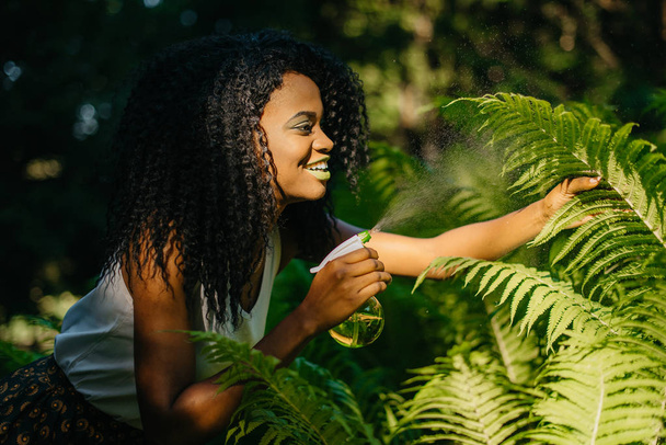 Attractive smiling african girl with green eye shadows and lipstick is using the green plastic trigger spray for ferns outdoor. - Photo, image