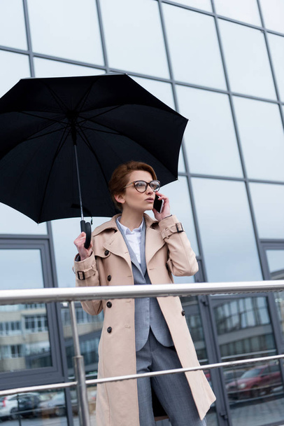 businesswoman with umbrella talking on smartphone on street - Zdjęcie, obraz