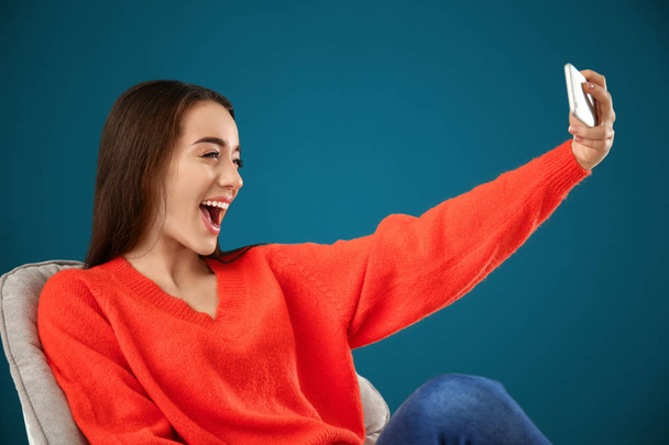 Happy young woman taking selfie while sitting in comfortable armchair against color background - Foto, Imagem