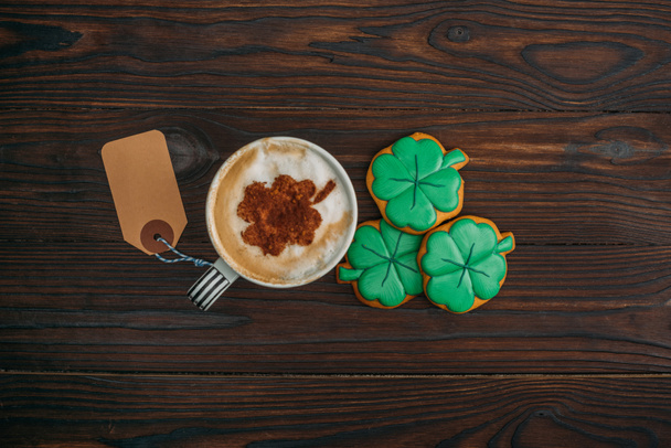 top view of cup of coffee with blank label and cookies in shape of clovers on wooden table - Photo, Image