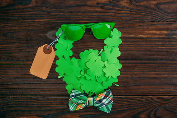 top view of bearded face symbol made of green clovers, sunglasses, blank tag and bow tie on wooden table - Foto, immagini