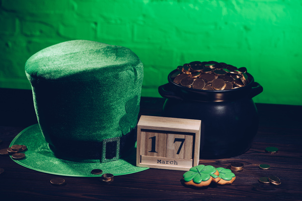 Ημερολόγιο, μπισκότα σε σχήμα τριφυλλιού, πράσινο ιρλανδικό καπέλο και δοχείο με χρυσά νομίσματα στο ξύλινο τραπέζι   - Φωτογραφία, εικόνα
