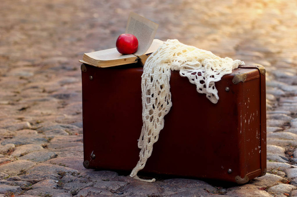 Manzana Roja, pañuelo blanco y libro viejo abierto en la vieja maleta de pie sobre los adoquines con manchas de sol al atardecer
 - Foto, Imagen