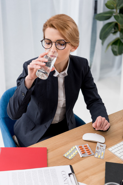 businesswoman in suit drinking water at workplace with pills in office - Foto, Bild