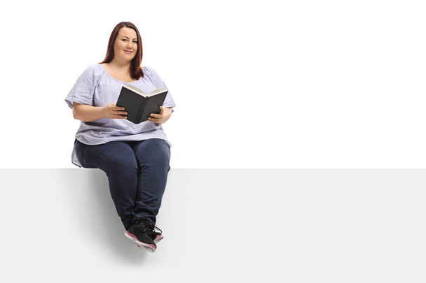Overweight woman with a book sitting on a panel and looking at the camera isolated on white background - Фото, зображення