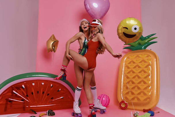 Full length of two attractive young women in swimwear embracing and smiling while standing against pink background - Fotografie, Obrázek