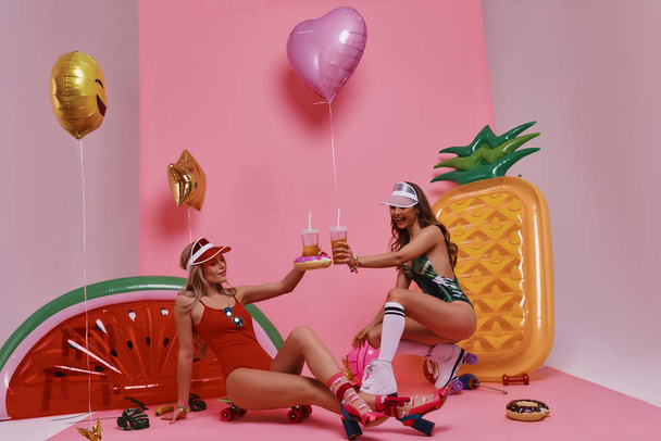 Beautiful young women in swimwear toasting each other and smiling while sitting against pink background - Foto, imagen