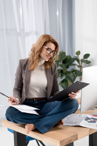 smiling businesswoman in eyeglasses doing paperwork while sitting on table in office - Foto, afbeelding