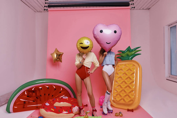 Full length of two women in swimwear covering faces with balloons while standing against pink background - Fotografie, Obrázek